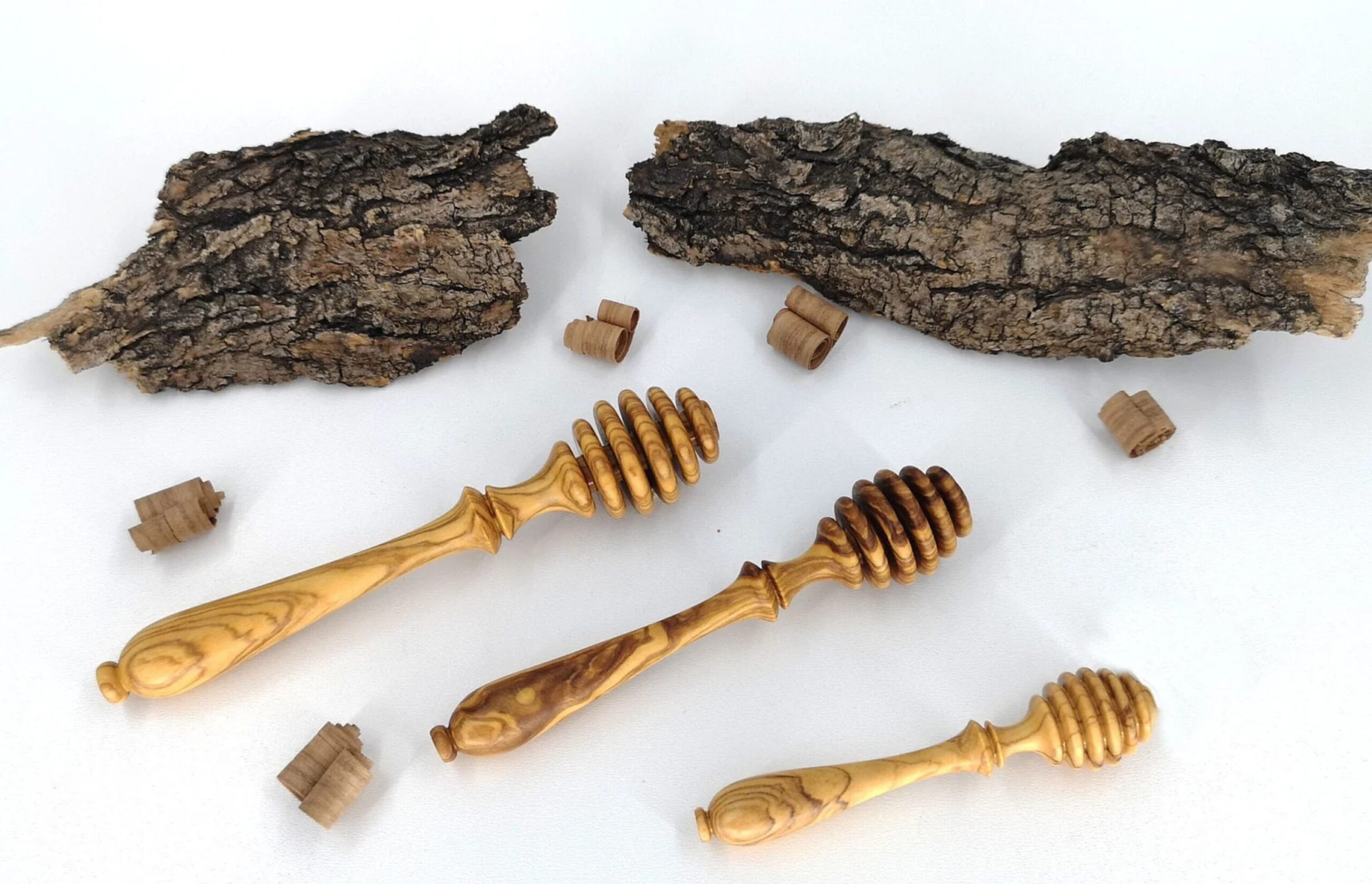 A close-up of three wooden honey dippers resting on a textured surface, accompanied by a few pieces of natural wood. The honey dippers have deep grooves and a smooth wooden finish, arranged neatly to showcase their design.