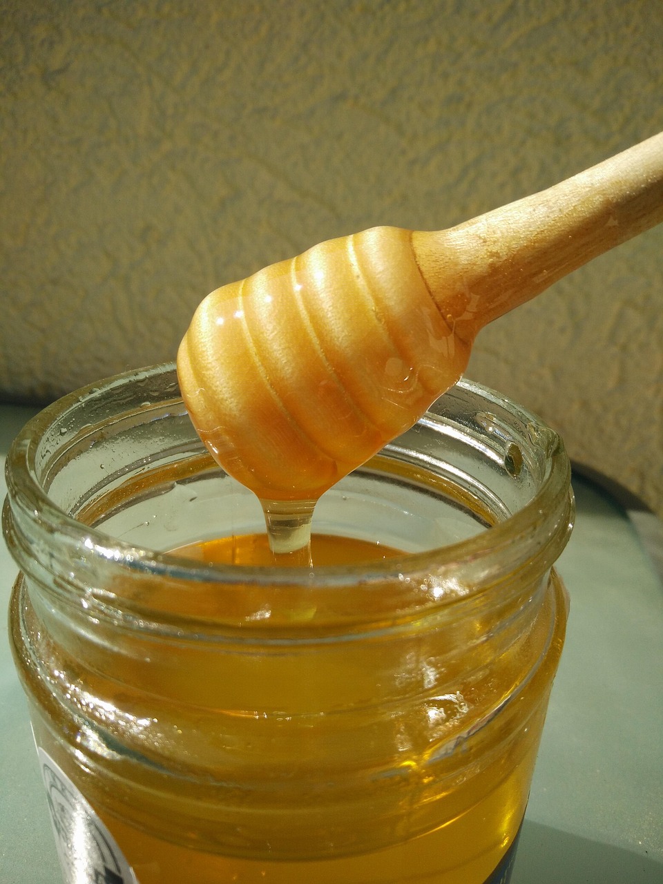 A glass jar filled with golden honey, with a wooden honey dipper coated in honey resting on top. The honey is glistening in the light, slowly dripping from the dipper. The background has a textured beige surface.