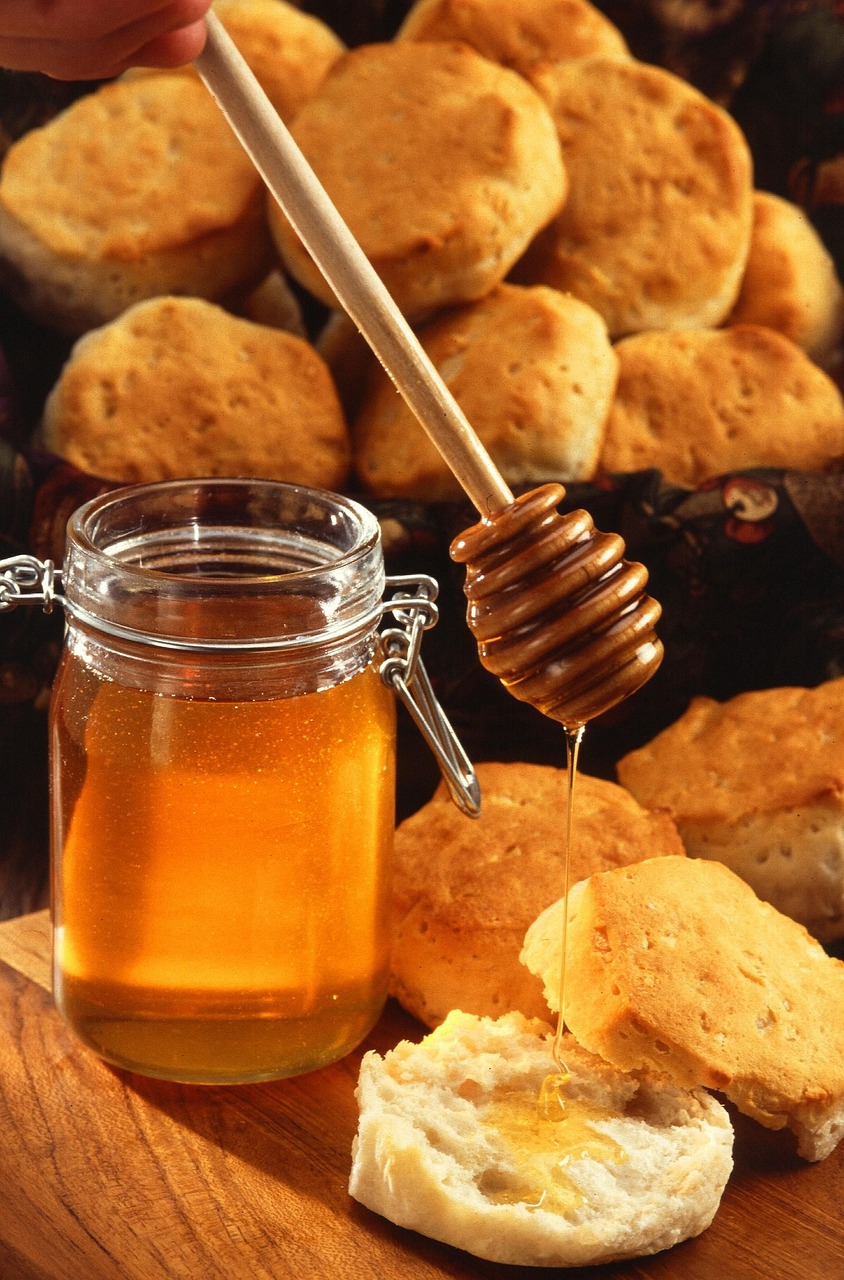 A glass jar of honey with a metal clasp, surrounded by freshly baked golden biscuits. A honey dipper is held above a biscuit, with honey drizzling onto the soft, fluffy interior. The setting has a warm, rustic feel.
